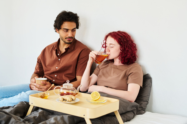 A dark-haired man holds a mug in his hands while sitting on a bed with a sick friend who drinks tea with lemon with a tray in front of her on which there is lemon ginger and honey