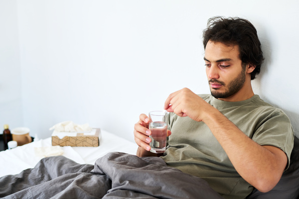 A guy with a cold sitting in bed under a gray blanket holding a glass of water in his hand throws a soluble tablet into it