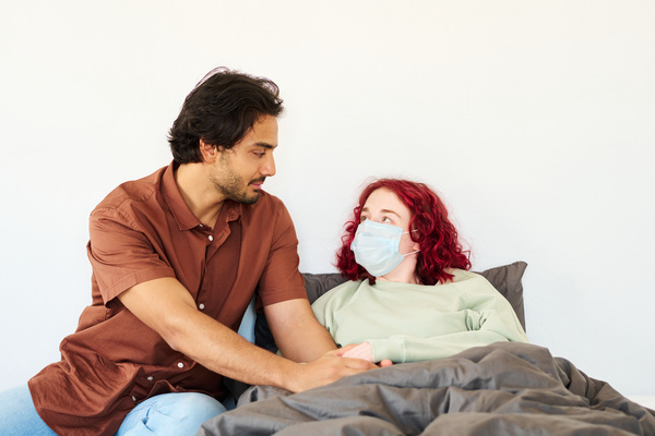A man with dark hair and beard dressed in a shirt holds the hands of a sick friend with red hair in a medical mask who is lying in a gray bed