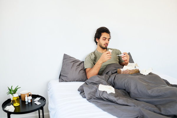 A young man with the flu is sitting in his pajamas in a gray bed with a box of paper napkins in front of him and holding a thermometer in his hand