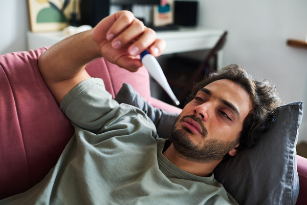 An unshaven guy with black tousled hair who is lying at home on the couch in pajamas and holding a thermometer with temperature readings has the flu