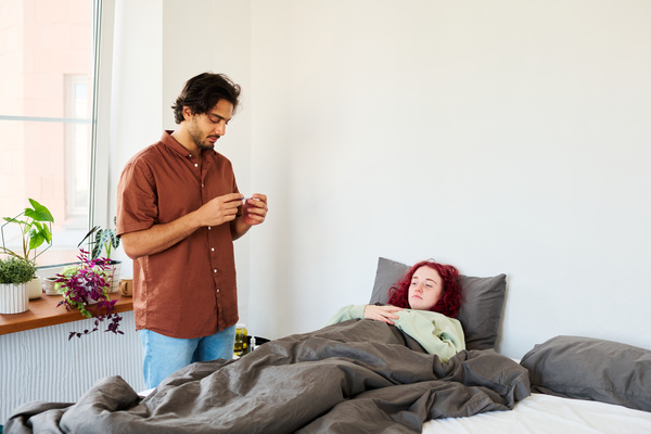 A dark-haired man in a shirt has measured measuring the temperature of his sick girlfriend with red hair lying in a gray bed looks at a thermometer that shows her temperature