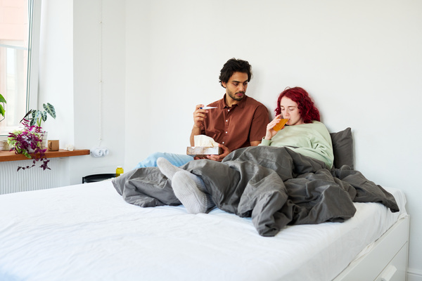 A dark-haired man with a beard in a brown shirt holds a thermometer and napkins while his sick girl with red hair dressed in pajamas is sitting on the bed drinking medicine