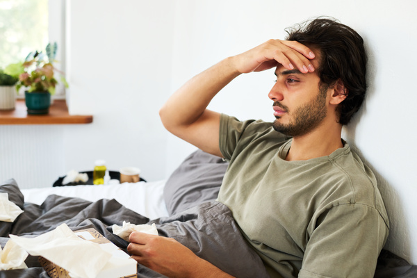 A guy with dark hair and beard has the flu and is lying in bed at home surrounded by paper napkins and holding his head