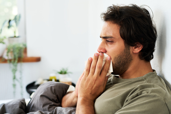 A young man has a sore throat and coughs while sitting at home in bed with a paper handkerchief pressed to his mouth
