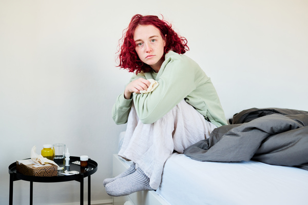 A sick woman with red curly hair dressed in home clothes is sitting with a melancholic look in the living room on the sofa with her arms wrapped around her knees and holding a paper handkerchief in her hand
