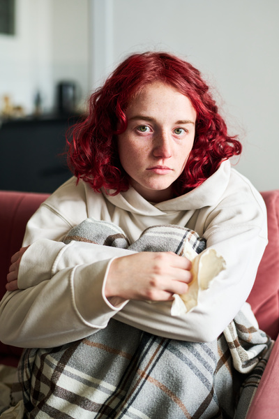 A red-haired girl with SARS dressed in light clothes with a depressive look is sitting on the sofa in the living room hugging her knees with her hands and holding a paper handkerchief in her hand