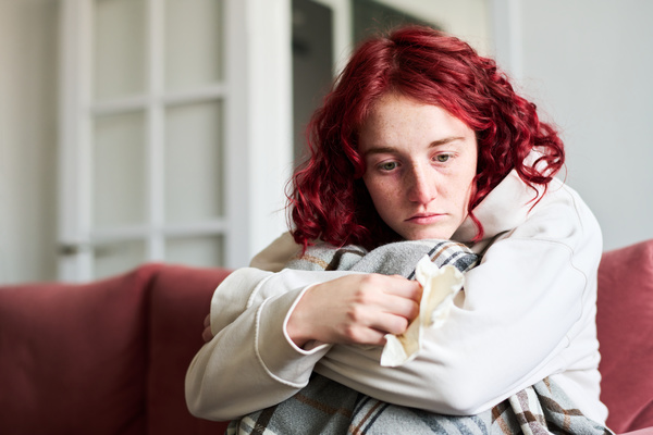 A female with flu with red curly hair dressed in home is sitting on the sofa with a melanccholic look hugging her knees with her hands and resting her head on them