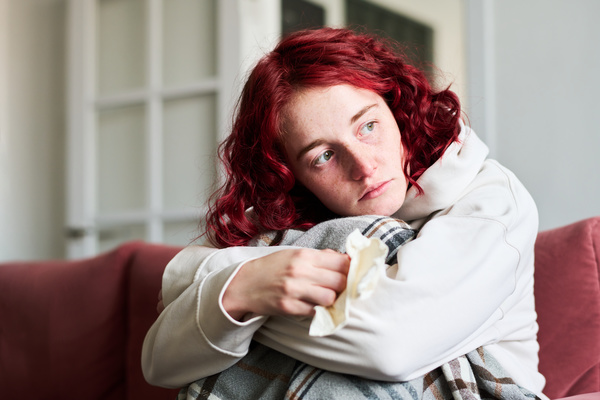 A woman with a cold with red curly hair dressed in a white hoodie is covered with a blanket and sits with a melancholic look on the sofa with her arms around her knees and her head on them