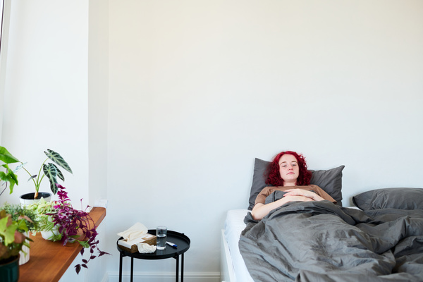 A girl with an illness with short red hair lying in bed folded her arms on her chest looks in front of her and is bored in a light room with a table with medicines and napkins next to a wooden window sill on which there are various flowers