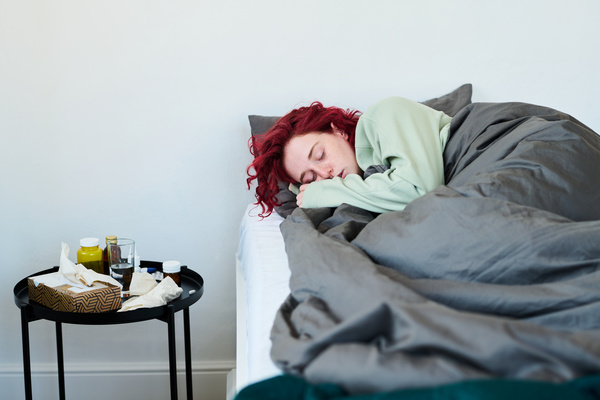 A young woman with curly red hair has a cold and sleeps on a bed covered with a gray blanket curled up on her side