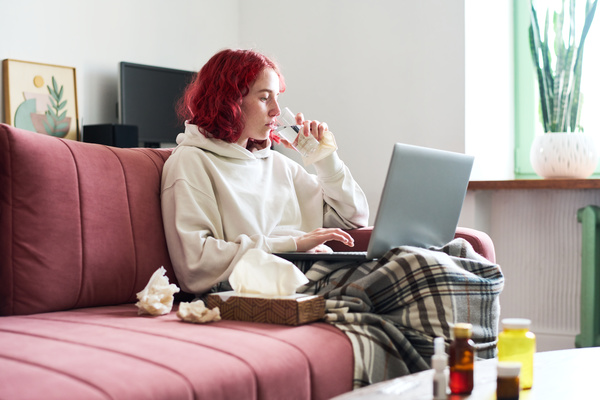 A woman with a runny nose sitting on a pink sofa with paper napkins drinks water and types on a laptop that is on her lap