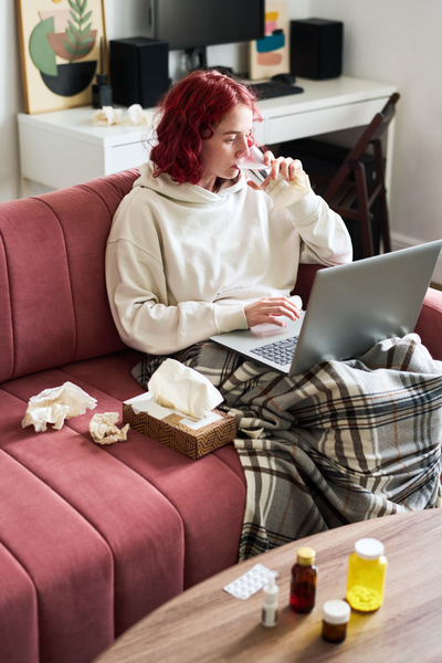 A sick woman with bright hair sitting on a pink sofa with paper handkerchiefs in a box and next to her drinking water and surfing the Internet on a silver laptop
