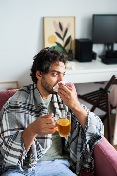A male with a runny nose is sitting on the sofa in the living room with a mug of tea with lemon covered with a blanket with a checkered pattern wiping his nose with a paper handkerchief