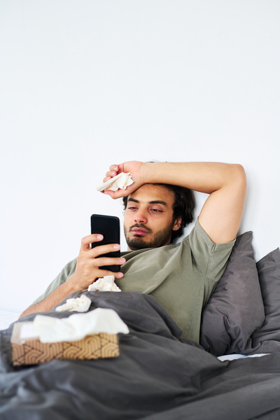 A man with black hair and stubble who has fallen ill with SARS is lying in a dark-colored bed with a box of paper handkerchiefs and surfing the Internet on his phone with one hand on his head
