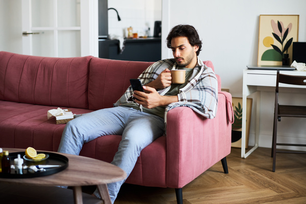 A guy with dark hair and beard who has SARS is sitting on a pink sofa with a plaid blanket on his shoulders and a mug of tea in his hand and watching a video on his phone