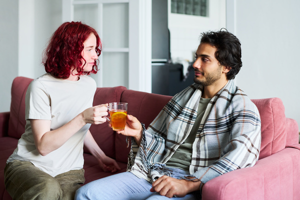 A girl with short red hair in a light T-shirt passes a mug of tea with lemon to a sick friend with dark hair and stubble who is wrapped in a plaid plaid sitting on a pink sofa in a bright living room