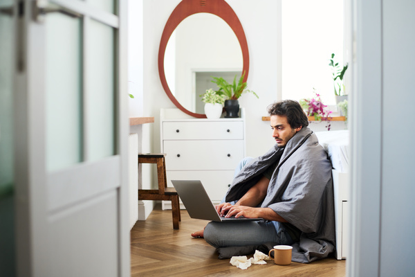 A guy with dark hair and stubble dressed in pajamas with a runny nose is sitting on the floor with a blanket covering his shoulders with a laptop on his lap and a mug of tea and crumpled paper handkerchiefs next to him