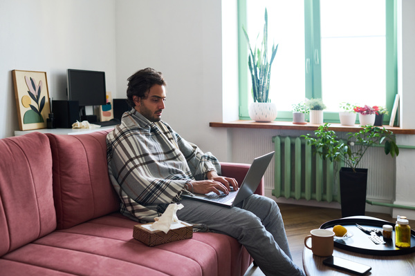 A sick man with dark hair and stubble is sitting on a pink sofa in the living room with a box of paper napkins next to him and a table with medicines covered with a plaid in a cage working on a laptop