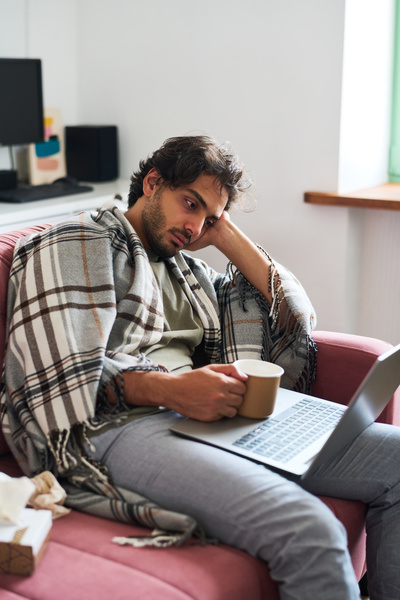 A man with uncombed hair got sick and sitting on the couch covering his shoulders with a blanket holds a mug in his hand and watches a movie on a laptop with a tired look