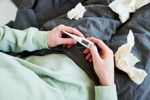 A person holds an electronic thermometer with high temperature readings while sitting in a pajamas in bed with paper handkerchiefs.