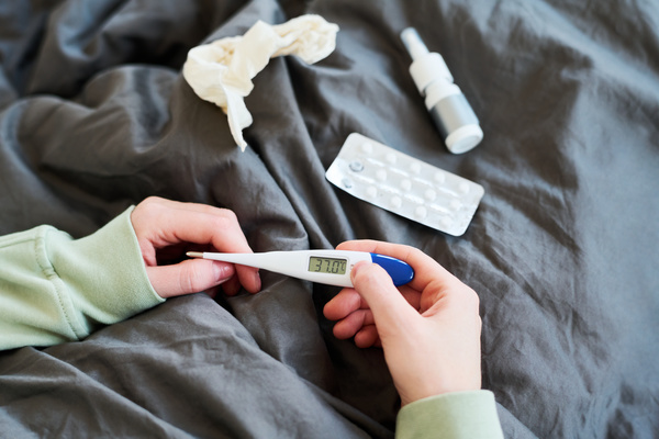 A person with a fever sitting in bed and covered with a gray blanket with pills a spray for a runny nose and a paper handkerchief and holding an electronic thermometer in hands