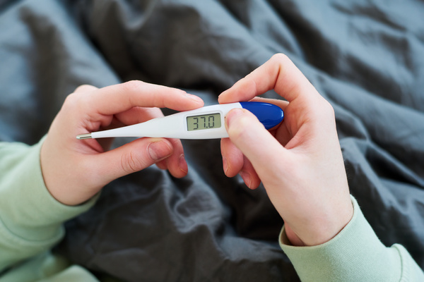 A person with a raised temperature holds an electronic thermometer with both hands showing the temperature value while sitting in bed in light pajamas