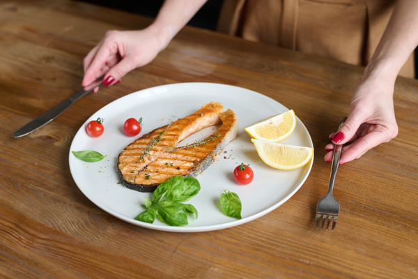 Grilled fish steak with thyme on a white platter with cherry basil leaves and lemon is on the table in front of a woman holding a fork and knife