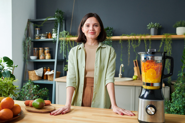 A smoothie maker with dark hair in a light green shirt stands with her hands on the table on which fruits are lying and there is a blender with ingredients for a drink in it