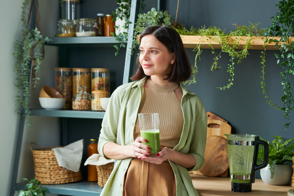 A woman with short dark hair dressed in a light green shirt over a beige top looking away holds a glass with a green smoothie in her hands