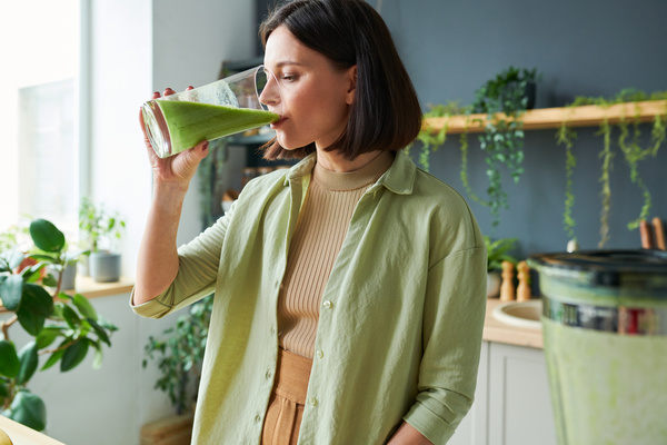 A woman with dark short-cropped hair wearing a light green shirt over a top standing in the kitchen drinking a green smoothie from a glass