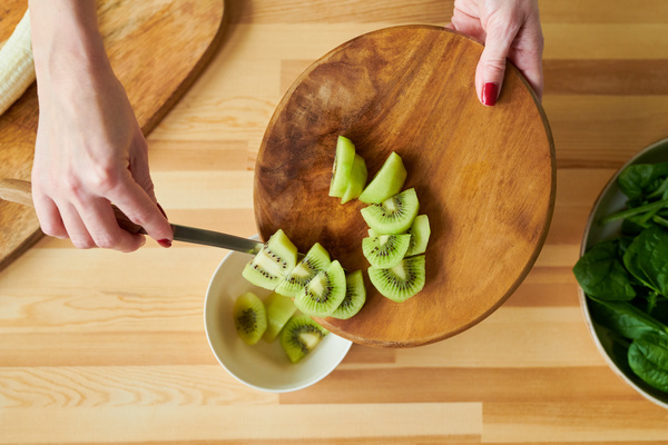 Smoothie maker  rakes peeled and diced kiwi with a knife from a wooden board into a white bowl