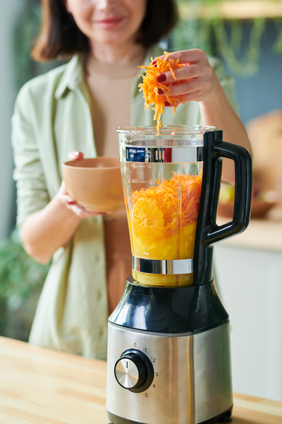 The smoothie maker puts grated carrots in a jug of a stationary blender with sliced orange that stands on a wooden table