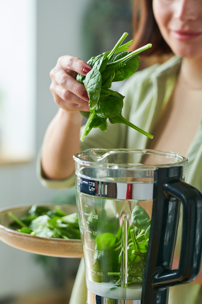 A woman making a smoothie puts greens from a light dish into a stationary blender