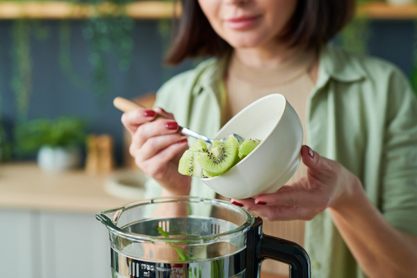 A woman in light clothes pours kiwi slices with a spoon from a white bowl into a blender jug