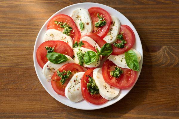 Caprese salad with pesto sauce and basil is neatly served on a white platter that is on a wooden table
