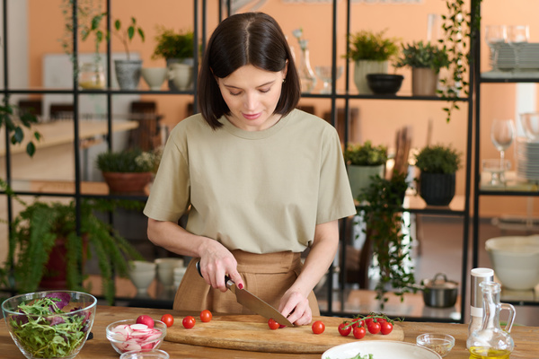 A female chef with dark loose hair at a table against the background of shelves with potted plants is slicing cherry tomatoes on a board next to which the ingredients are laid out in bowls