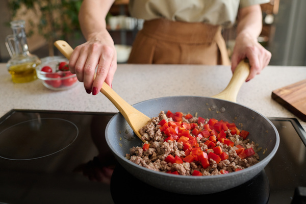 A cook in light clothes is frying minced meat with chopped red bell pepper stirring it in a frying pan with a chef's spoon