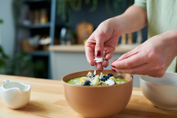 A woman sprinkles coconut chips on a green smoothie with pieces of fruit and blueberries in a sand-colored bowl standing on a wooden table next to a white gravy boat and a bowl