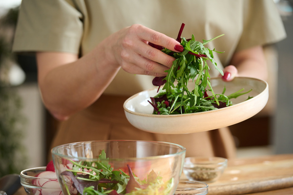 A chef with wine-tinged nails in a beige outfit puts arugula and basil on a white platter  which she holds over a glass bowl with salad greens