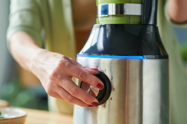 A woman with a wine-colored manicure turns on a stationary silver-black blender on the table to make a smothie