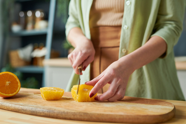 A smoothie maker in light-colored clothes standing at the kitchen table on a wooden cutting board cuts a peeled orange