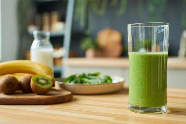 A glass with a green smoothie is on a table made of light wood against a background of fruit
