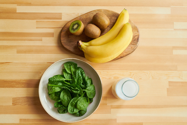 Spinach leaves on a platter bananas and kiwi on a cutting board and milk in a glass jar on a wooden table