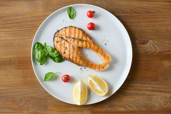 A dish consisting of red fish steak with basil cherry tomatoes and lemon sliced on a white platter