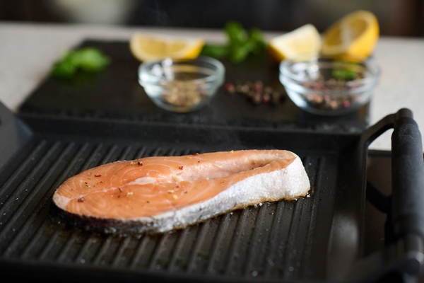 A piece of red fish on a bone is being cooked on a black grill with seasonings and lemon slices lying on the board behind