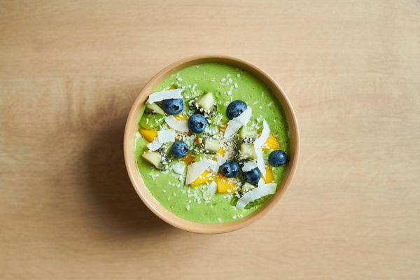 A beige bowl with a green smoothie with mango kiwi and blueberries sprinkled with coconut chips is on a wooden surface