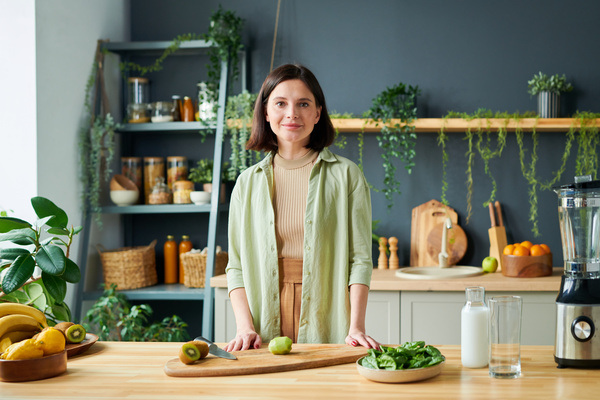 A woman who makes smoothies is standing at a table on which kiwis with a knife are lying on a cutting board and there is a dish of spinach and a jug of milk next to it