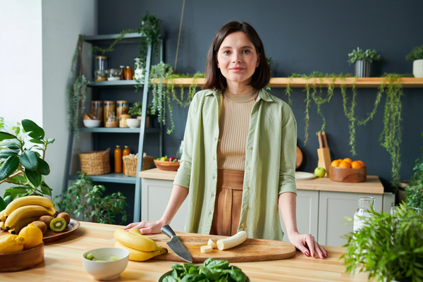 Portrait of a smoothie maker with dark short hair in light clothes standing in the kitchen at a table on which there is a knife with a sliced banana on the board and fruits and greens in the plates