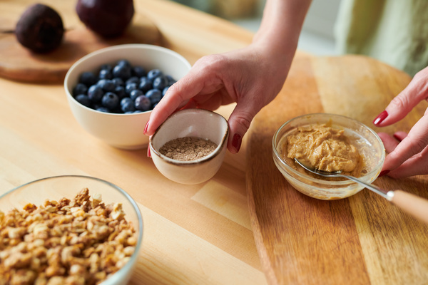 A woman takes a small bowl with vegetable protein powder from a table on which there is peanut paste in a bowl with a spoon  a plate with blueberries and granola
